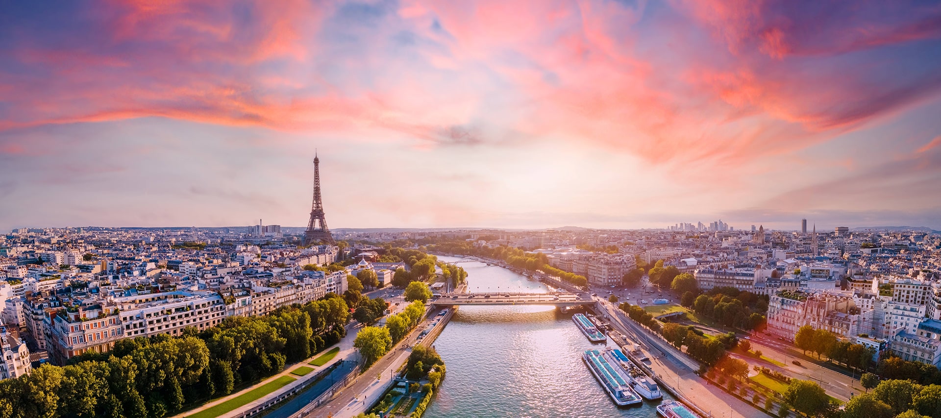 Antiquaire et brocanteur à Paris Vue panoramique de Paris au coucher du soleil. La Tour Eiffel se dresse bien en évidence sur la gauche, tandis que la Seine serpente à travers la ville remplie de bateaux. L'horizon est parsemé de bâtiments et le ciel est peint de teintes vibrantes de roses et d'oranges, ajoutant un accueil chaleureux au paysage.