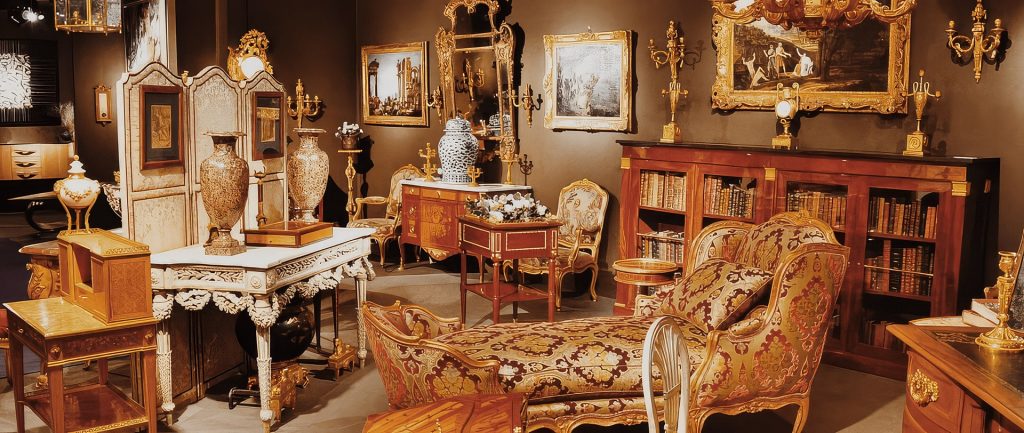 Antiquaire et brocanteur à Paris Une salle antique richement décorée offre un accueil chaleureux avec ses meubles ornés et ses œuvres d'art. L'espace comprend un bureau en bois finement sculpté, une chaise longue avec un revêtement floral, une variété de vases et des appliques murales dorées. Des peintures dans des cadres ornés et des étagères remplies de livres embellissent les murs sombres.