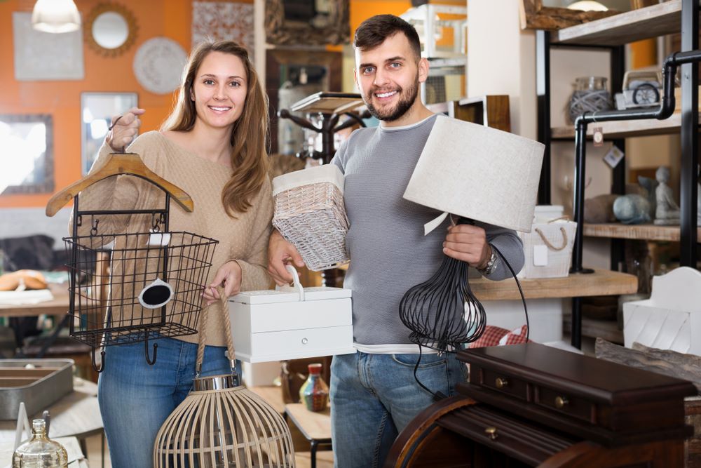 Antiquaire et brocanteur à Paris Une femme et un homme souriants se tiennent debout dans un magasin d’articles pour la maison, tenant dans leurs mains divers objets de décoration qu’ils viennent de choisir dans le cadre de leur sélection Accueil. La femme tient une cage à oiseaux en fil métallique, une lampe en bois et un petit meuble à tiroirs blanc. L'homme tient un panier en osier et une lampe en métal avec un abat-jour beige. Des étagères avec des objets de décoration sont visibles en arrière-plan.