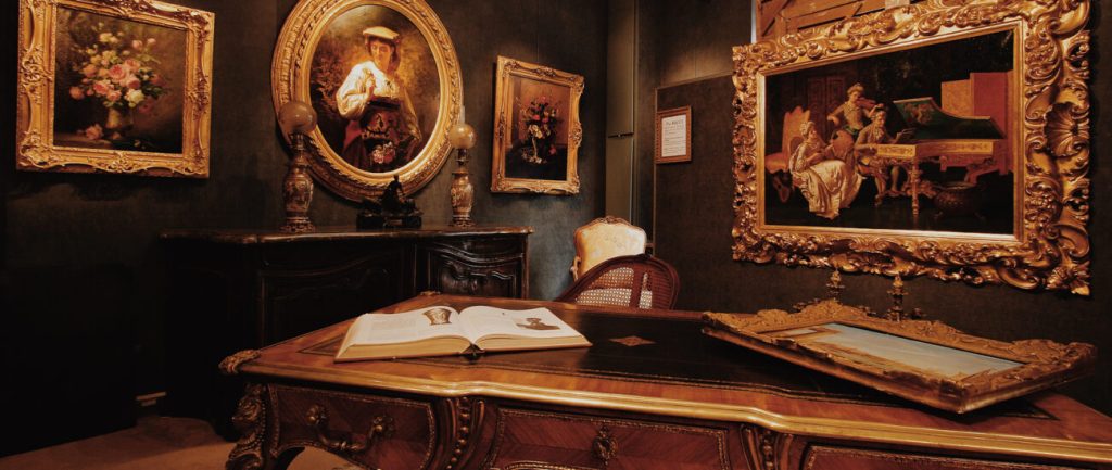 Antiquaire et brocanteur à Paris Une pièce opulente aux murs sombres présente des peintures encadrées ornées, des meubles anciens complexes et des objets décoratifs. Un grand bureau en bois au premier plan présente un livre ouvert et un tableau encadré. Des chaises élégantes et un grand meuble complètent cet accueillant luxueux.