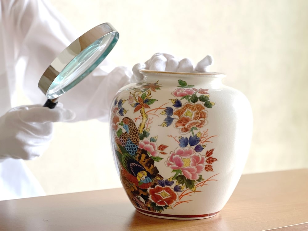 Antiquaire et brocanteur à Paris Une personne portant des gants blancs tient une loupe près d’un vase décoratif en porcelaine, qui présente des motifs complexes et colorés de fleurs et d’un paon. La scène suggère une estimation ou une appréciation de l’œuvre d’art sur un fond uni et clair.