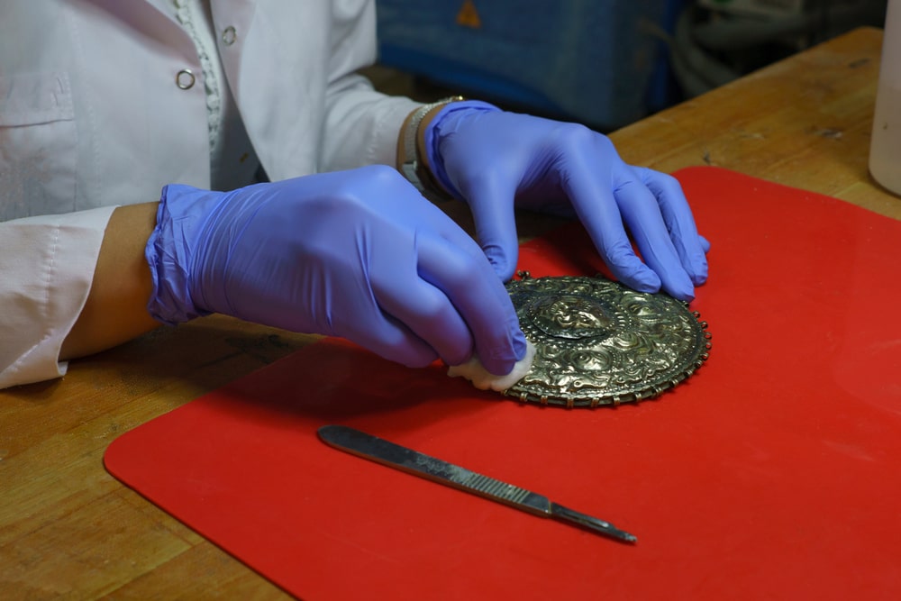 Antiquaire et brocanteur à Paris Une personne portant une blouse blanche et des gants bleus nettoie méticuleusement un artefact métallique de conception complexe avec un chiffon blanc sur un tapis rouge. Un outil semblable à un scalpel se trouve à côté d’eux sur la table en bois. Le décor semble être celui d’un atelier ou d’un laboratoire de restauration, où une estimation précise des détails est primordiale.