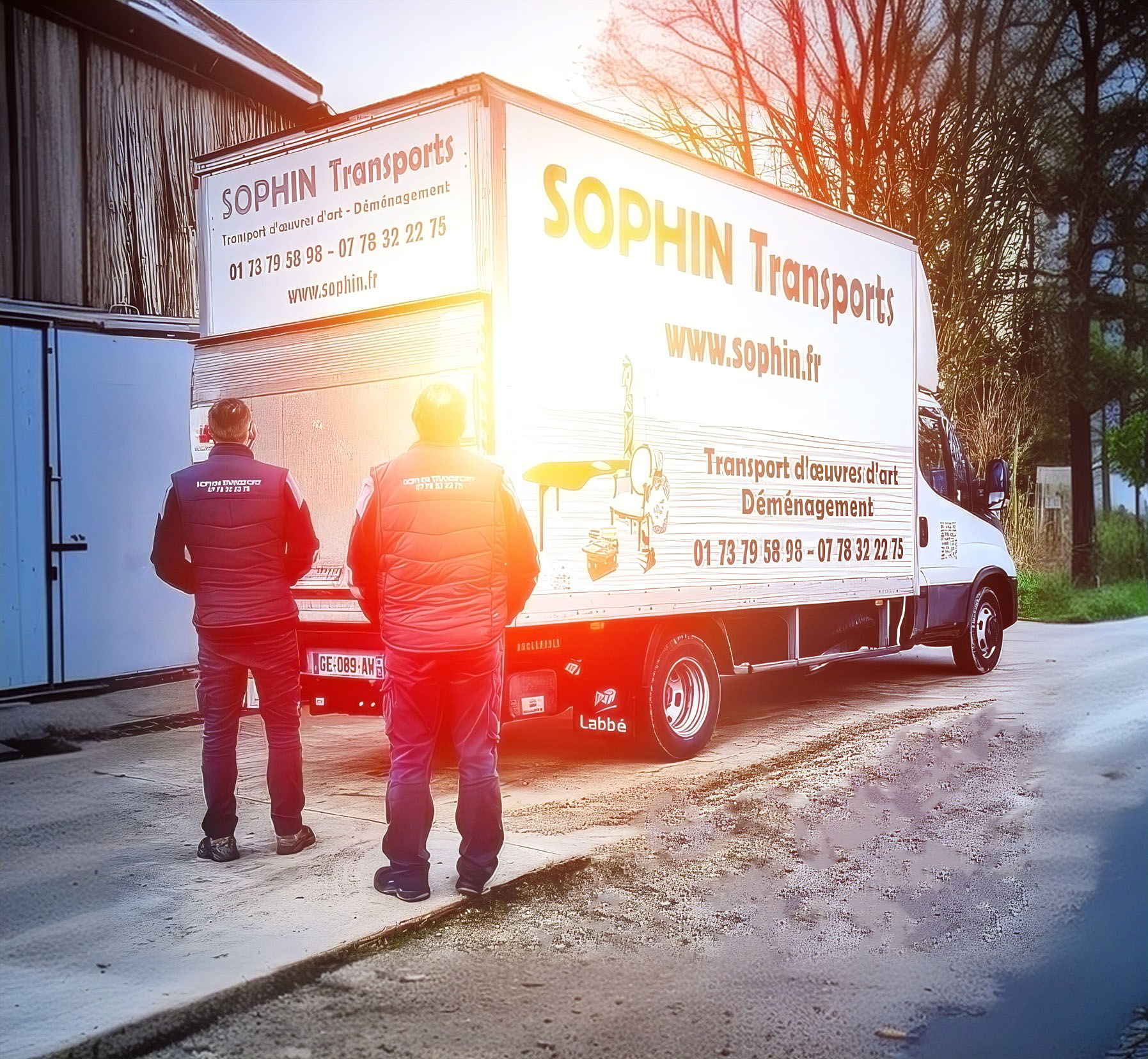 Deux personnes en uniformes de travail se tiennent devant un camion de transport d'œuvres d'art "SOPHIN Transports", stationné près d'un bâtiment.