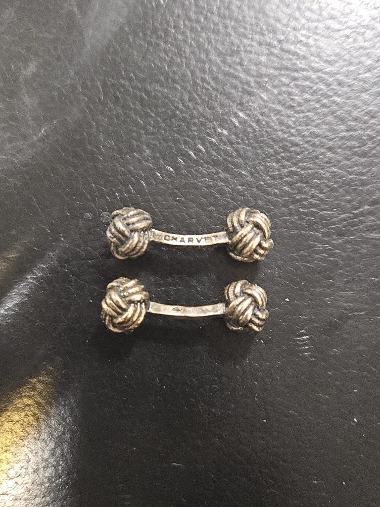 Antiquaire et brocanteur à Paris Une paire de boutons de manchette argentés repose sur une surface en cuir noir. Chaque bouton de manchette comporte une barre métallique incurvée reliant deux extrémités minutieusement nouées. Le mot "CHARYT" est gravé sur l'une des barres de liaison, faisant allusion à ses origines parisiennes de La Maison Chavet. La surface du cuir a une texture légèrement brillante.