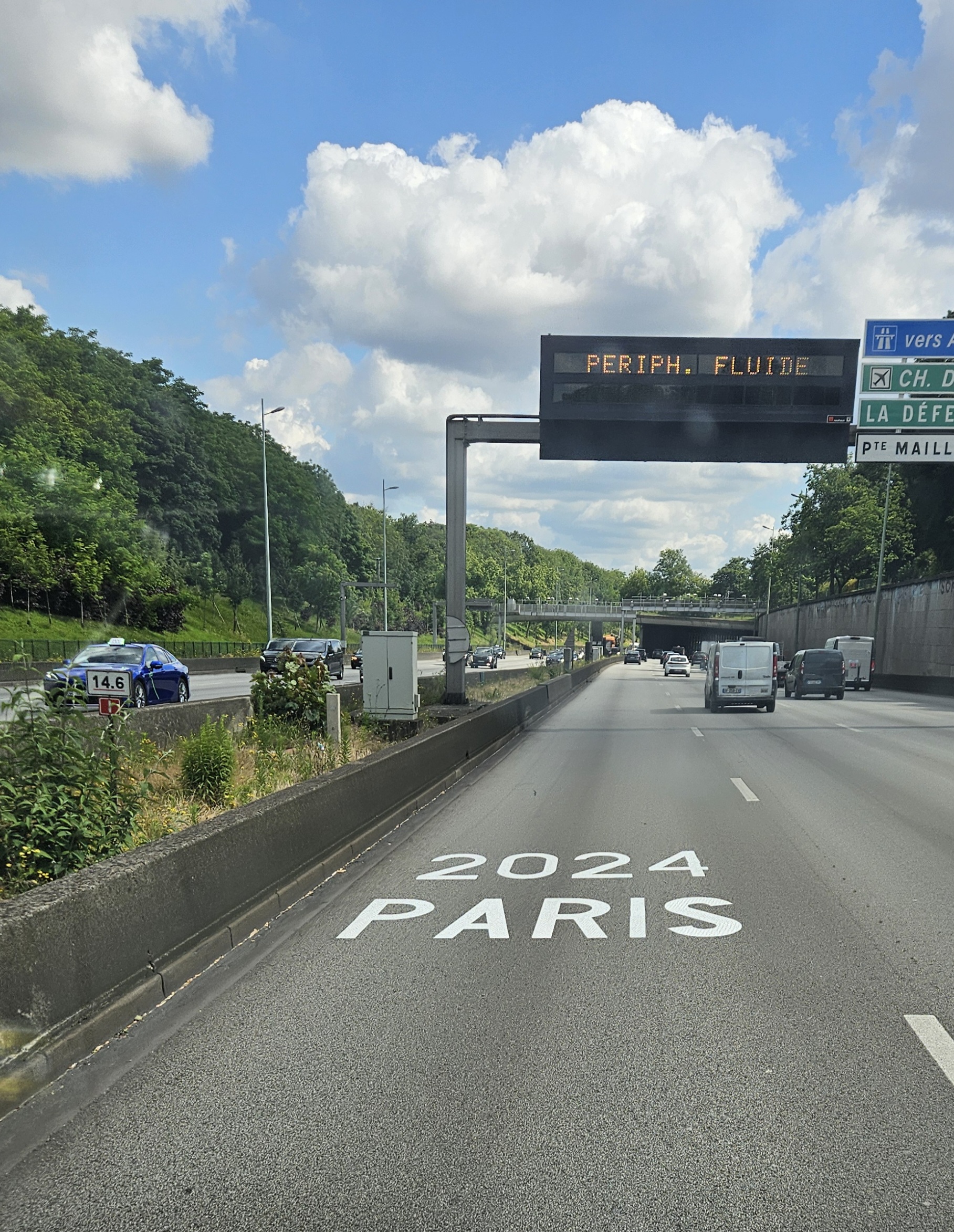 Antiquaire et brocanteur à Paris Une scène d'autoroute avec plusieurs voies et des véhicules circulant dans les deux sens. Un panneau numérique affiche « PERIPH. FLUIDE », indiquant des conditions de circulation fluides. La route est balisée « 2024 PARIS JO ». Des arbres et du feuillage bordent les deux côtés de l’autoroute sous un ciel partiellement nuageux.