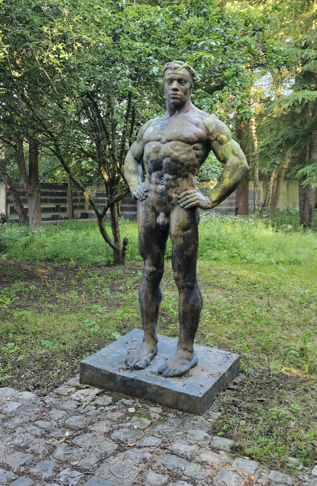 Antiquaire et brocanteur à Paris Une statue en bronze patiné représentant un homme nu debout, les mains sur les hanches, est située dans un cadre extérieur verdoyant et luxuriant à Paris. La statue est montée sur un socle en pierre ; le sol autour est pavé et herbeux. L’arrière-plan présente des arbres et une zone herbeuse. Estimation gratuite disponible.