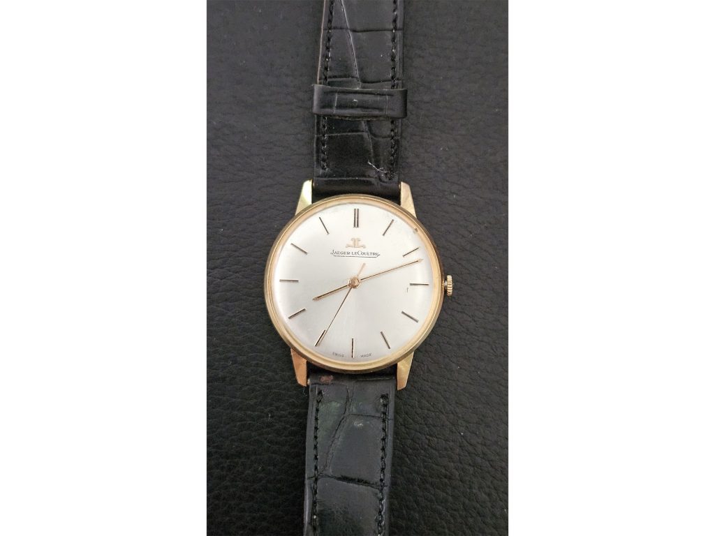 Antiquaire et brocanteur à Paris Une montre-bracelet sophistiquée avec un boîtier rond doré, un cadran blanc et de fins index noirs. La montre a des aiguilles noires élégantes et porte la marque « Jaeger-LeCoultre » sous la position 12 heures. Parfait pour ceux qui s'intéressent à l'achat de montres, il est doté d'un bracelet en cuir noir texturé avec une boucle.