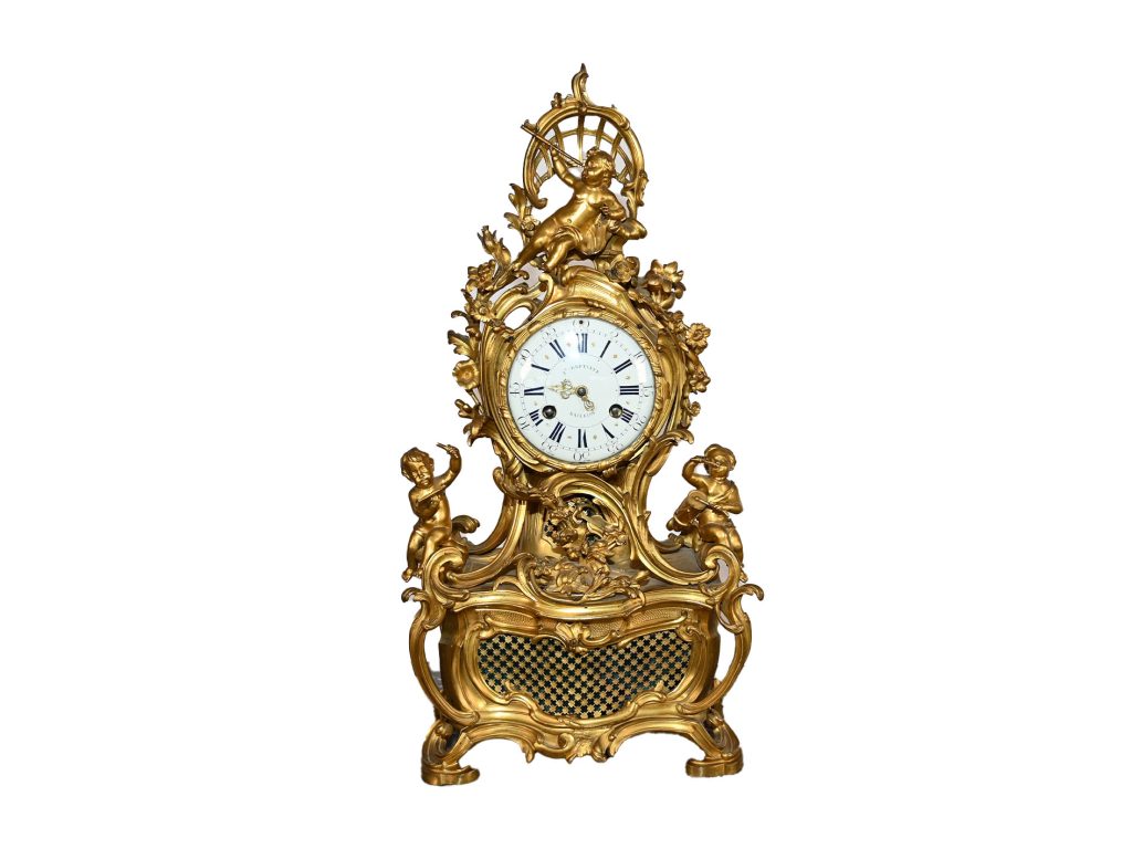 Antiquaire et brocanteur à Paris Une horloge antique ornée d'un cadran blanc et de chiffres romains est enfermée dans un cadre doré élaboré décoré de motifs complexes et de figures de chérubins. La partie inférieure présente un motif en damier, tandis que la partie supérieure présente un chérubin supplémentaire assis sous une structure en forme de treillis, incarnant L'Héritage Intemporel de Joseph de Saint-Germain.