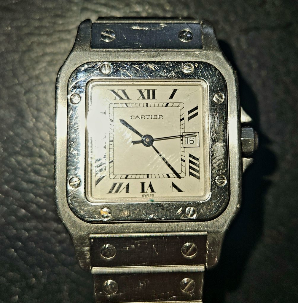 Antiquaire et brocanteur à Paris Découverte : Montre-bracelet à cadran carré avec bracelet métallique affiché. Le cadran de la montre comporte des chiffres romains noirs et un guichet de date à 3 heures. Le nom de la marque « Cartier » est visible sur le cadran, indiquant ce modèle de Montre Santos. Il présente une finition distinctive en métal brossé, rehaussée de vis sur la lunette.